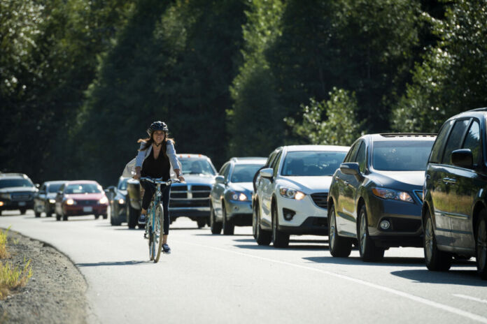 Eine glücklich aussehende Frau fährt Fahrrad auf der Straße. Neben ihr stehen viele Autos im Stau, während sie an ihnen vorbei pendelt und sie überholt. Dabei trägt sie einen Helm und lächelt.