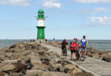 Einige Fahrradfahrer in professioneller Ausstattung stehen an der steinigen Küste der Ostsee. Im Hintergrund prangt ein weiß-grüner Leuchtturm, unter dem weitere Touristen stehen. Das Meer ist ruhig und weit.