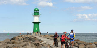 Einige Fahrradfahrer in professioneller Ausstattung stehen an der steinigen Küste der Ostsee. Im Hintergrund prangt ein weiß-grüner Leuchtturm, unter dem weitere Touristen stehen. Das Meer ist ruhig und weit.