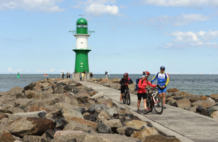 Einige Fahrradfahrer in professioneller Ausstattung stehen an der steinigen Küste der Ostsee. Im Hintergrund prangt ein weiß-grüner Leuchtturm, unter dem weitere Touristen stehen. Das Meer ist ruhig und weit.