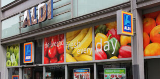 Blick auf eine moderne Aldi-Filiale. Im Eingangsbereich sind die oberen Scheiben mit Bildern von Obst und Gemüse beklebt. Im unteren Bereich der Scheiben befindet sich Glaskästen mit den aktuellen Angeboten der Woche.