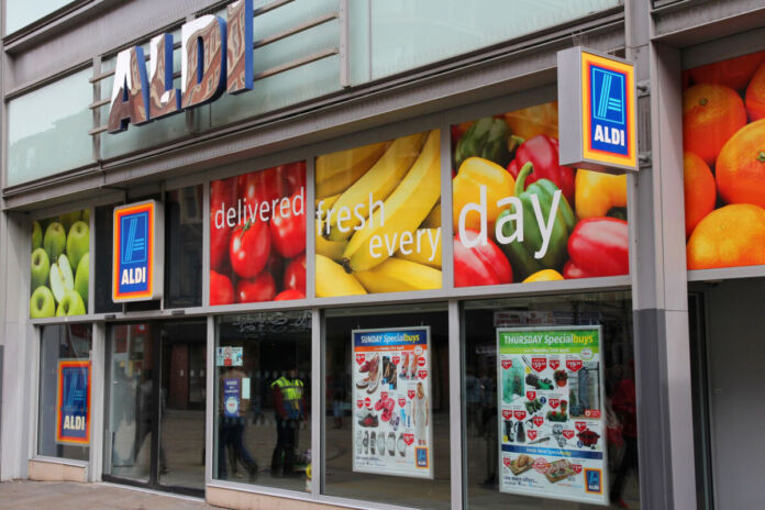 Blick auf eine moderne Aldi-Filiale. Im Eingangsbereich sind die oberen Scheiben mit Bildern von Obst und Gemüse beklebt. Im unteren Bereich der Scheiben befindet sich Glaskästen mit den aktuellen Angeboten der Woche.