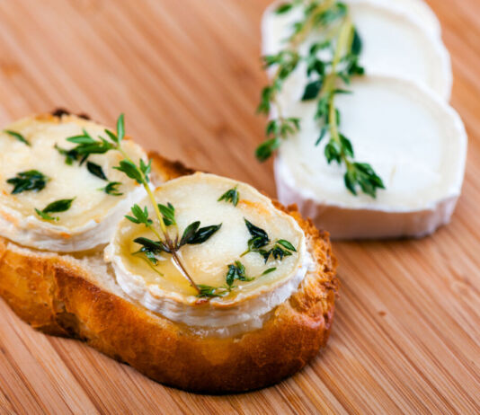 Auf einem Holzbrett liegt eine Scheibe Brot, die mit zwei Scheiben Ziegenkäse belegt ist. Darauf befindet sich gratinierter Honig und Thymian. Neben dem Brot liegen zwei weitere Ziegenkäse-Scheiben, die mit einem Thymianzweig dekoriert sind.