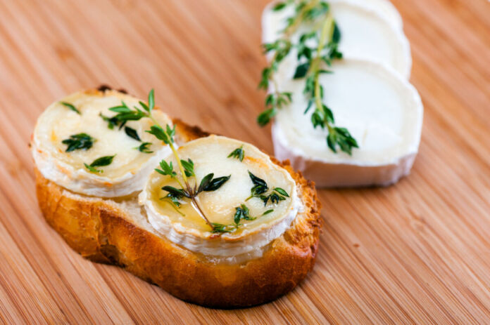 Auf einem Holzbrett liegt eine Scheibe Brot, die mit zwei Scheiben Ziegenkäse belegt ist. Darauf befindet sich gratinierter Honig und Thymian. Neben dem Brot liegen zwei weitere Ziegenkäse-Scheiben, die mit einem Thymianzweig dekoriert sind.