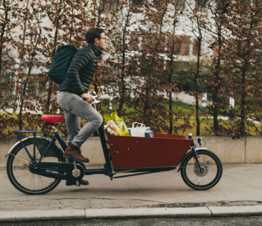 Ein Mann sitzt auf einem Lastenfahrrad und scheint einige Einkäufe an Kunden auszuliefern. Ein Lastenfahrrad unterscheidet sich von einem normalen Fahrrad durch einen vorne angebrachten Kasten, in dem man zum Beispiel seine Einkäufe verstauen kann.