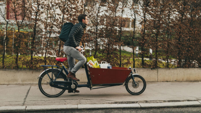 Ein Mann sitzt auf einem Lastenfahrrad und scheint einige Einkäufe an Kunden auszuliefern. Ein Lastenfahrrad unterscheidet sich von einem normalen Fahrrad durch einen vorne angebrachten Kasten, in dem man zum Beispiel seine Einkäufe verstauen kann.