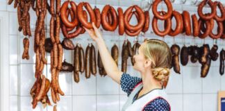 Verkäuferin in einer Metzgerei greift hinter der Theke nach oben zu einer geräucherten Wurst, die an einer Wand mit weißen Fliesen hängt. Es sind unterschiedliche Wurstwaren zu sehen.