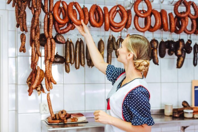 Verkäuferin in einer Metzgerei greift hinter der Theke nach oben zu einer geräucherten Wurst, die an einer Wand mit weißen Fliesen hängt. Es sind unterschiedliche Wurstwaren zu sehen.