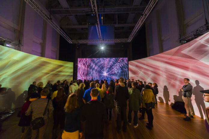 Zahlreiche Menschen besuchen eine Show oder eine Ausstellung, die mit Illumination und Lichtern arbeitet. Es ist dunkel und die verschiedenen LEDs scheinen auf eine große Leinwand.