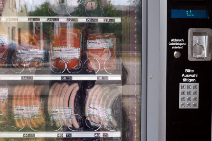 In einem Automaten werden verschiedene Grillspezialitäten angeboten. Hinter der Glasscheibe befinden sich etliche Würste und diverses Grillfleisch. Der Automat spart Personalkosten ein.