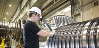 Ein Facharbeiter in der Fabrik bei der Arbeit. Er trägt Schutzbrille und Schutzhelm und hantiert an einer großen Maschine aus Stahl oder Metall. Die Fabrik für Maschinenbau ist riesig.