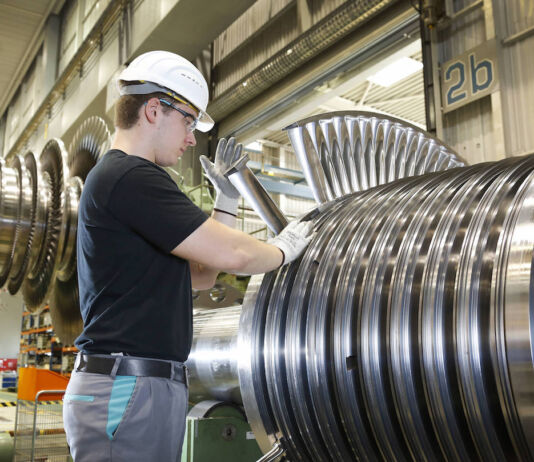 Ein Facharbeiter in der Fabrik bei der Arbeit. Er trägt Schutzbrille und Schutzhelm und hantiert an einer großen Maschine aus Stahl oder Metall. Die Fabrik für Maschinenbau ist riesig.