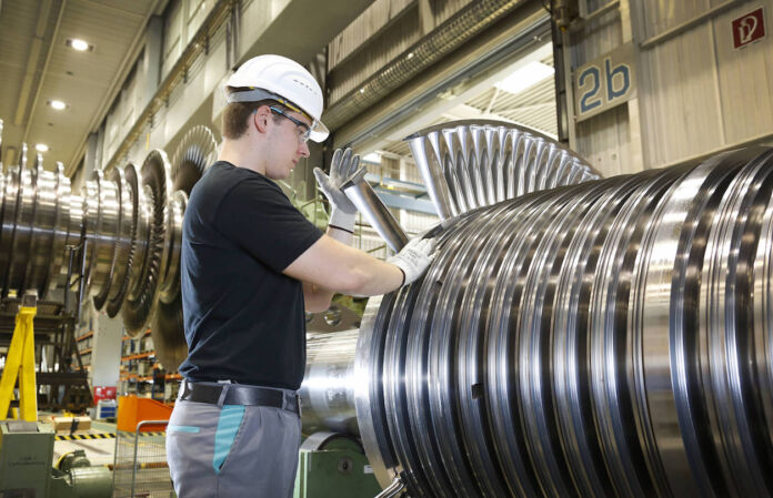 Ein Facharbeiter in der Fabrik bei der Arbeit. Er trägt Schutzbrille und Schutzhelm und hantiert an einer großen Maschine aus Stahl oder Metall. Die Fabrik für Maschinenbau ist riesig.