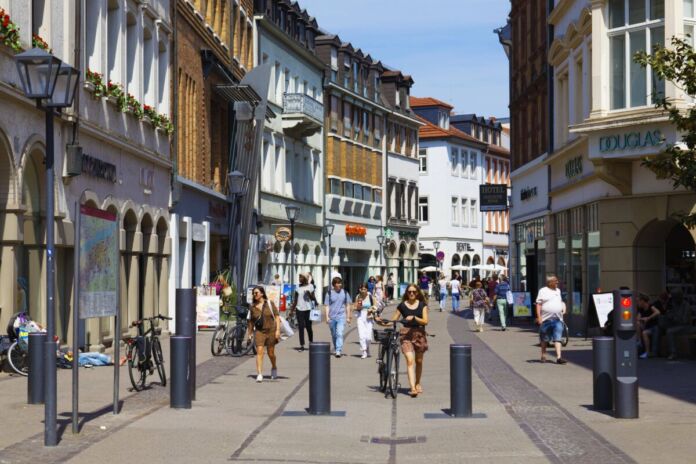 In einer Fußgängerzone in einer Altstadt schlendern mehrere Fußgänger durch die Innenstadt. Sie gehen an einigen Einkaufsläden vorbei. Einfahrtsperren verhindern, dass Autos durch die Innenstadt fahren können.