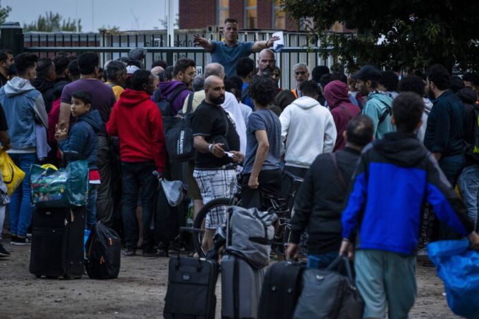 Zahlreiche Flüchtlinge stehen mit gepackten Koffern und Taschen vor einer Unterkunft. Sie suchen ein neues Zuhause und stehen dafür lange Zeit in einer Schlange.