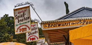 An einer Bude auf einem Volksfest gibt es Bratwurst, Pommes und andere Speisen und Getränke. Am Dach hängen Banner, auf denen verschiedene Gerichte stehen.