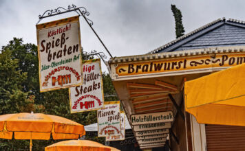 An einer Bude auf einem Volksfest gibt es Bratwurst, Pommes und andere Speisen und Getränke. Am Dach hängen Banner, auf denen verschiedene Gerichte stehen.