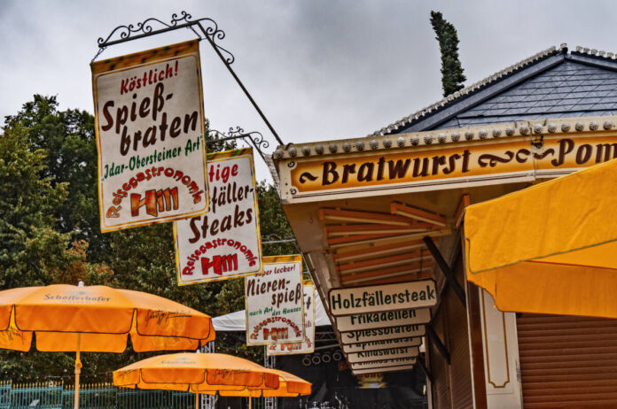 An einer Bude auf einem Volksfest gibt es Bratwurst, Pommes und andere Speisen und Getränke. Am Dach hängen Banner, auf denen verschiedene Gerichte stehen.