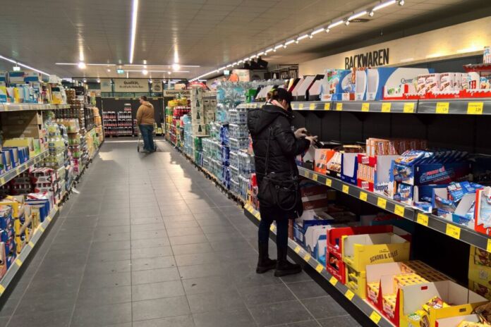 In einem Supermarkt oder Discounter steht eine Kundin vor den Regalen mit Süßigkeiten und greift nach etwas. Weiter hinten steht ein Kunde bei den Getränken.