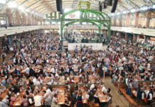 Eine große Menschenmenge feiert gemeinsam in einem Festzelt das Oktoberfest. Sie tragen Tracht oder normale Kleidung und genießen die Spezialitäten wie Bier und zünftigen Schweinsbraten.