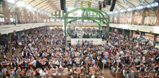 Eine große Menschenmenge feiert gemeinsam in einem Festzelt das Oktoberfest. Sie tragen Tracht oder normale Kleidung und genießen die Spezialitäten wie Bier und zünftigen Schweinsbraten.