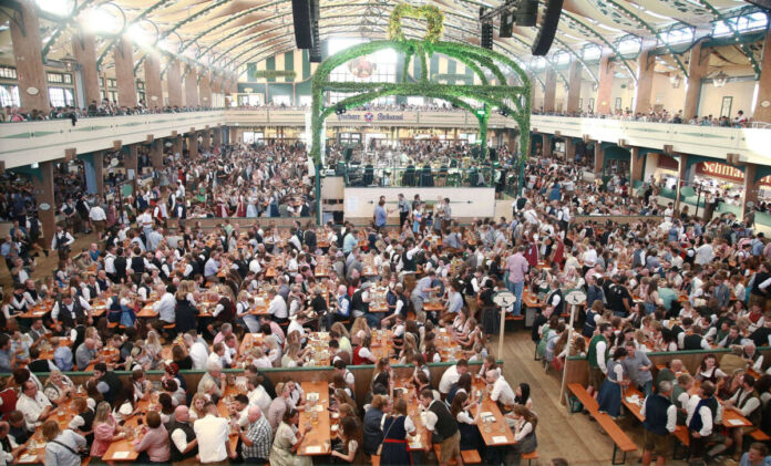 Eine große Menschenmenge feiert gemeinsam in einem Festzelt das Oktoberfest. Sie tragen Tracht oder normale Kleidung und genießen die Spezialitäten wie Bier und zünftigen Schweinsbraten.