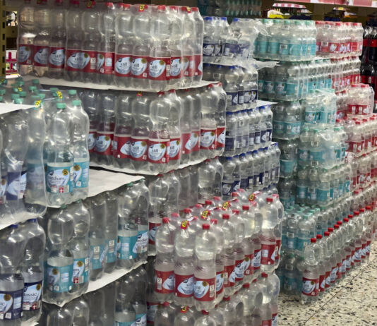 Gestapeltes Wasser in Plastikflaschen in einem Supermarkt. Zur Auswahl steht stilles Wasser und Wasser mit Kohlensäure. Im Hintergrund stehen neben dem Wasser auch Plastikflaschen mit Cola.