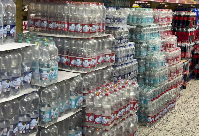 Gestapeltes Wasser in Plastikflaschen in einem Supermarkt. Zur Auswahl steht stilles Wasser und Wasser mit Kohlensäure. Im Hintergrund stehen neben dem Wasser auch Plastikflaschen mit Cola.