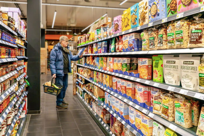 Ein Kunde befindet sich im Supermarkt und hält eine Packung in der Hand. Er betrachtet das Müsli-Regal. Darin sind viele Sorten mit Müsli und Cornflakes ausgestellt. Auf der anderen Seite des Ganges befinden sich die Konserven in Gläsern und Dosen.