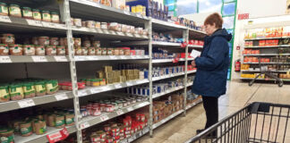 Eine nachdenkliche Dame im Supermarkt beim Einkaufen steht vor einem Regal mit verschiedenen Konserven und sucht nach dem richtigen Produkt. Sie hält eine Einkaufsliste in der Hand. Im Vordergrund steht ein Einkaufswagen.