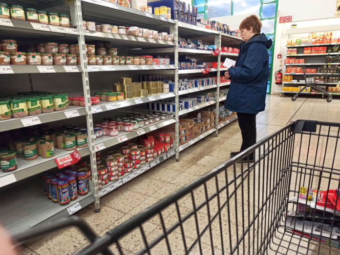 Eine nachdenkliche Dame im Supermarkt beim Einkaufen steht vor einem Regal mit verschiedenen Konserven und sucht nach dem richtigen Produkt. Sie hält eine Einkaufsliste in der Hand. Im Vordergrund steht ein Einkaufswagen.