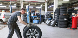 Ein Mann rollt einen Reifen durch eine Kfz-Werkstatt. Er wird die Autoreifen von einem Fahrzeug wechseln und sie deshalb zum Auto hinbewegen.