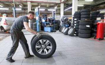 Ein Mann rollt einen Reifen durch eine Kfz-Werkstatt. Er wird die Autoreifen von einem Fahrzeug wechseln und sie deshalb zum Auto hinbewegen.