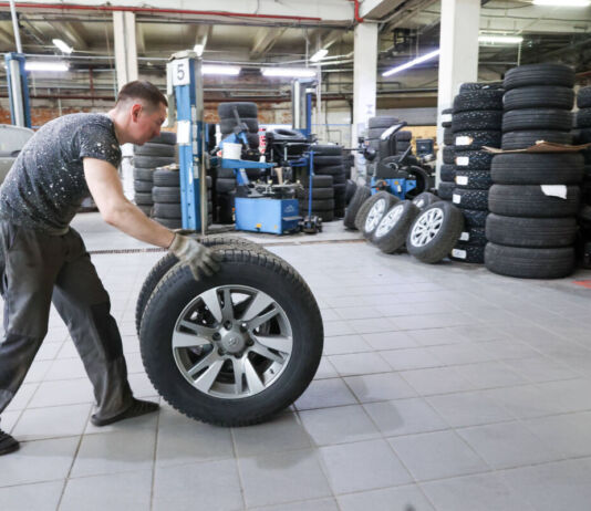 Ein Mann rollt einen Reifen durch eine Kfz-Werkstatt. Er wird die Autoreifen von einem Fahrzeug wechseln und sie deshalb zum Auto hinbewegen.