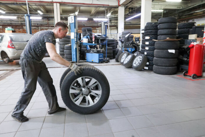 Ein Mann rollt einen Reifen durch eine Kfz-Werkstatt. Er wird die Autoreifen von einem Fahrzeug wechseln und sie deshalb zum Auto hinbewegen.