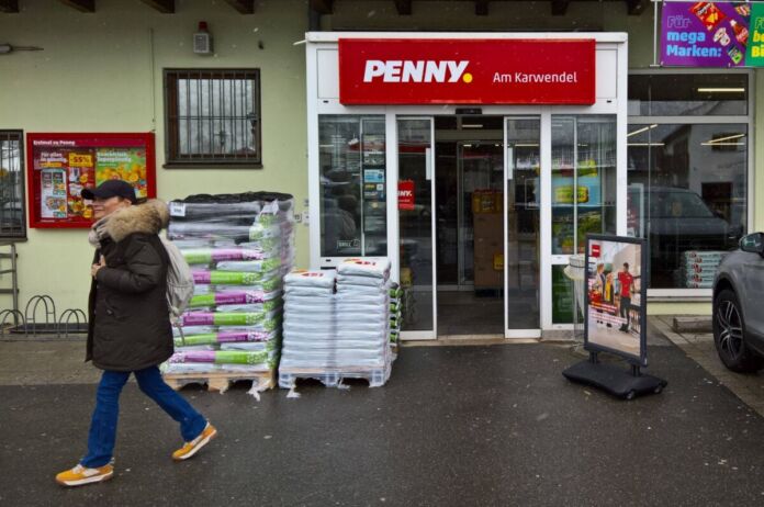 Der Eingangsbereich des bekannten Supermarkts Penny ist zu sehen. Ein Fußgänger läuft an dem Discounter vorbei. Vor dem Eingang stehen einige Produkte der Kette.