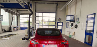 Ein Tesla Model 3 in roter Farbe steht in der Werkstatt, weil das E-Auto eine Reparatur braucht oder bei der Amtlichen Fahrzeuguntersuchung ist. Die Werkstatt-Halle ist groß und leer mit blauen Türen.