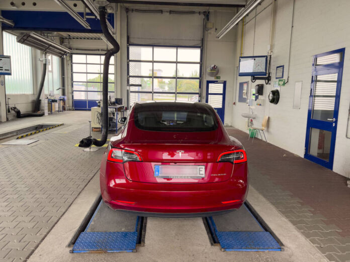 Ein Tesla Model 3 in roter Farbe steht in der Werkstatt, weil das E-Auto eine Reparatur braucht oder bei der Amtlichen Fahrzeuguntersuchung ist. Die Werkstatt-Halle ist groß und leer mit blauen Türen.