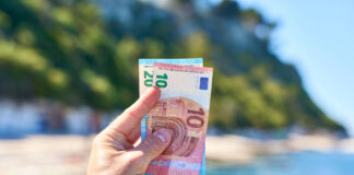 Eine männliche Hand hält Geld ins Bild, einen 10-Euro-Schein und einen 20-Euro-Schein. Im Hintergrund ist ein Urlaubsort mit Sonnenschein, Meer, Strand und schöner Natur.