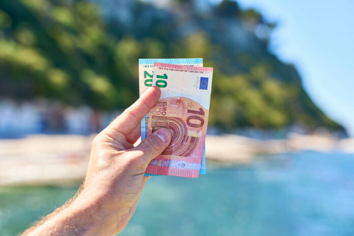 Eine männliche Hand hält Geld ins Bild, einen 10-Euro-Schein und einen 20-Euro-Schein. Im Hintergrund ist ein Urlaubsort mit Sonnenschein, Meer, Strand und schöner Natur.