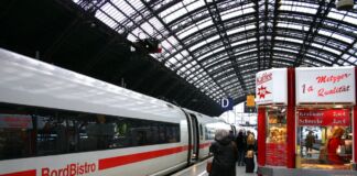 Ein Zug wartet auf dem Bahnsteig auf Fahrgäste. Auf dem Zug ist die Beschriftung "Bordbistro" zu lesen. Vor dem Zug stehen Fahrgäste. Am Bahnsteig gibt es einen Imbiss.