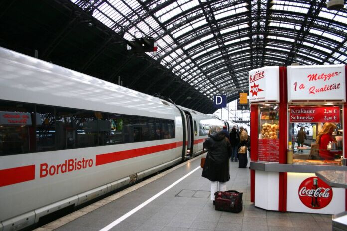 Ein Zug wartet auf dem Bahnsteig auf Fahrgäste. Auf dem Zug ist die Beschriftung "Bordbistro" zu lesen. Vor dem Zug stehen Fahrgäste. Am Bahnsteig gibt es einen Imbiss.