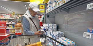 Eine ältere Frau steht vor einem Regal mit Mehlpackungen und hält eine Packung Mehl in der Hand. Neben ihr steht ein Einkaufswagen, der schon mit Nahrungsmitteln wie Keksen, Brot, Möhren und Milch gefüllt ist.