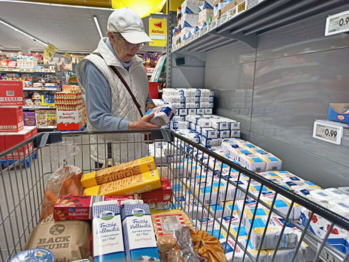 Eine ältere Frau steht vor einem Regal mit Mehlpackungen und hält eine Packung Mehl in der Hand. Neben ihr steht ein Einkaufswagen, der schon mit Nahrungsmitteln wie Keksen, Brot, Möhren und Milch gefüllt ist.