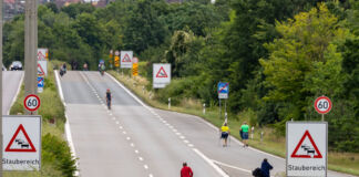 Eine breite Straße ist leer und wird nur mit zwei Fahrradfahrern befahren. An der Seite befinden sich Warnschilder und Baustellen-Zeichen. Die Straße neigt sich nach oben und es gilt Tempo 60.