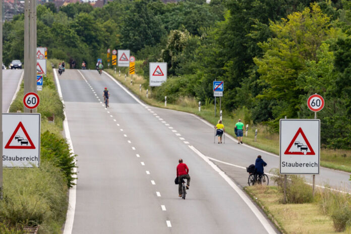 Eine breite Straße ist leer und wird nur mit zwei Fahrradfahrern befahren. An der Seite befinden sich Warnschilder und Baustellen-Zeichen. Die Straße neigt sich nach oben und es gilt Tempo 60.