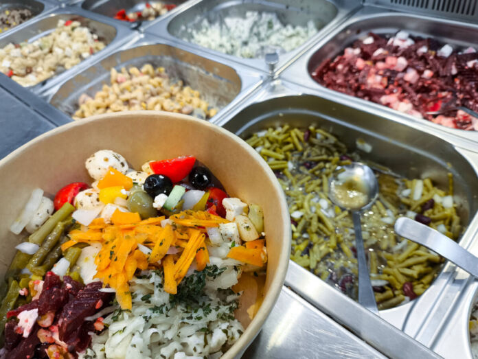 Theke in einem Supermarkt mit einer Auswahl an frischen Salaten und Gerichten zum Zusammenstellen und Mitnehmen. Die Selbstbedienungstheke bietet grüne Bohnen, Rotkohl, Oliven, Nudeln, Feta und gemischte Salatvariationen.