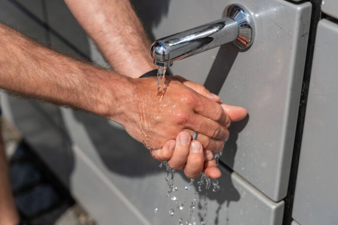 Ein Mann wäscht sich seine Hände unter laufendem Wasser, das aus einem Wasserhahn kommt. Das Wasser stammt aus einem öffentlichen Wasserspender.