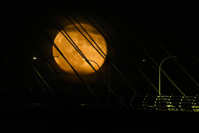 Vor einem pechschwarzen Himmel mitten in der Nacht sieht man einen vollen leuchtend orangefarbenen Mond. Vor ihm sind Stromseile und Laternen zu sehen. Es erwartet uns bald ein großes Himmelsspektakel über Deutschland.