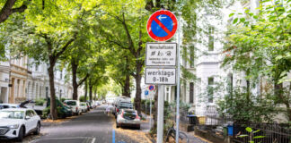 In einer Straße, die auf beiden Seiten von Häusern und Bäumen umsäumt ist, befindet sich eine blaue durchgezogene Linie. In blauer Schrift steht an der Linie Wirtschaftszone.